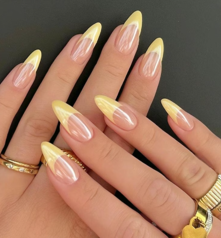 Close-up of hands with long almond-shaped nails featuring a pink-to-yellow gradient design, accessorized with gold rings.