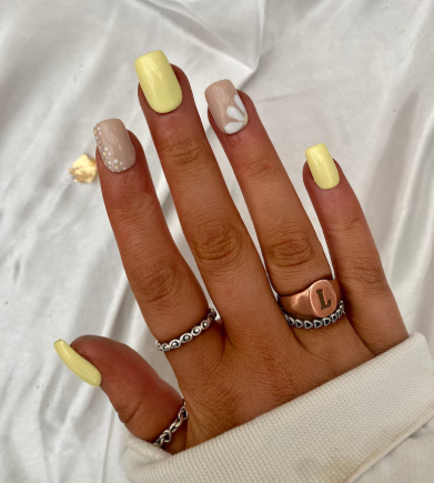 Hand with manicured nails featuring yellow polish on thumb, ring, and pinky; index nail with beige base and white polka dots; middle nail with beige base and white flower design. Adorned with silver and copper rings, including one with the letter “L,” set against white fabric.