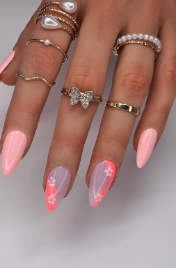 Close-up of a hand with long almond-shaped nails painted in pastel pink and lavender, featuring white floral designs and pink dots on two nails. The hand is adorned with multiple gold rings, including a pearl, a rhinestone butterfly, and other decorative styles.
