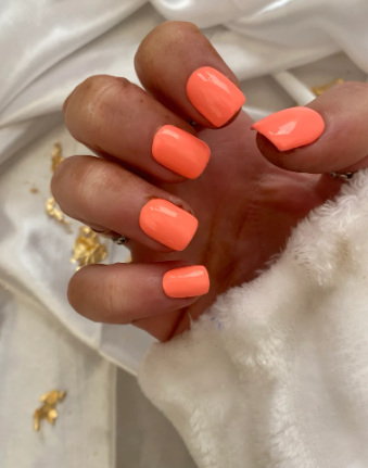 Bright orange summer manicure with glossy finish, holding white fluffy fabric, with gold flakes in the background.