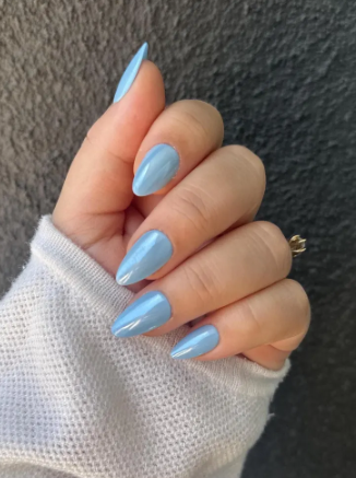 Glossy light blue almond-shaped nails with a gold ring, partially wrapped in a white sleeve, set against a dark textured background.