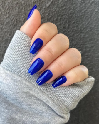 Glossy cobalt blue summer nails peeking from a light gray sweater sleeve, set against a textured gray background.