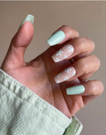 Close-up of a hand with mint green nails; index and middle fingers feature clear bases with white floral designs, while the other nails are solid mint green. The hand is partially wrapped in light green fabric
