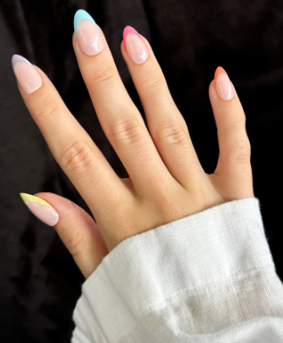 Hand with a white sleeve, showcasing manicured nails with a clear base and multicolored gradient tips—blue, pink, orange, yellow, and purple.