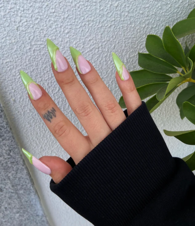 Hand with long, pointed nails featuring a light green and pink design, wearing a black sleeve, with a finger tattoo, set against a white textured wall and green leafy background.