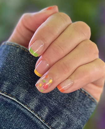 Close-up of a hand with a denim sleeve, featuring a clear base manicure with multicolored tips—green, yellow, orange, and red—and a thumb decorated with colorful polka dots.