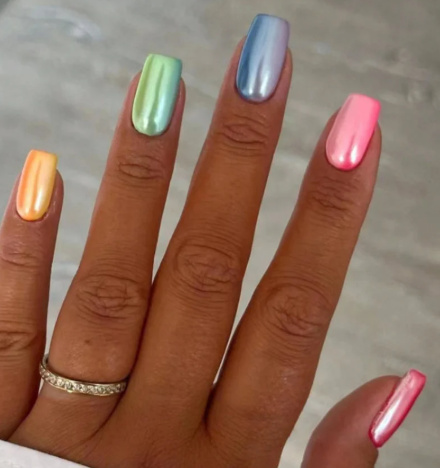lose-up of a hand with multicolored metallic nails—orange, green, blue, pink, and red—wearing a gold ring with small diamonds, against a neutral blurred background