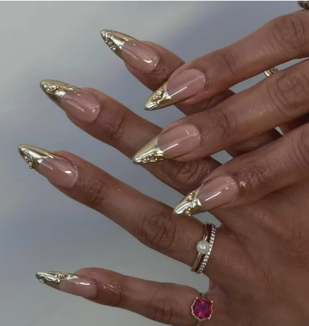 Close-up of hands with long pointed nails featuring a nude base and gold metallic tips, adorned with rings including a pearl and a large pink gemstone.