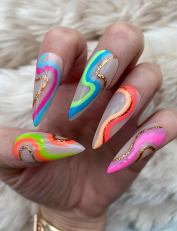 Close-up of a hand with long pointed acrylic nails featuring neon wavy designs in pink, orange, green, blue, and yellow, accented with gold glitter, set against a soft white fabric background.