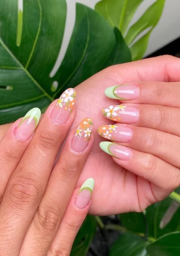 Close-up of hands with manicured nails featuring green French tips and floral designs with white flowers and yellow centers on a light orange base, set against large tropical green leaves.